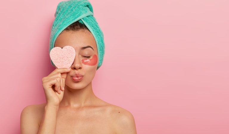 Isolated shot of pretty female model has eyes closed, lips rounded, stands with bare shoulders, enjoys beauty treatments, holds cosmetic sponge over eye, cares about face wears hydrogel patches, towel